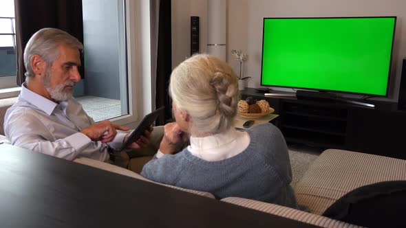 Elderly Couple Sits on A Couch in A Living Room, Watches TV with A Green Screen and Work on Tablet