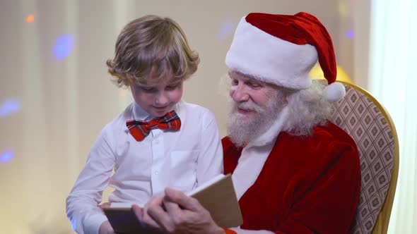 Portrait of Happy Boy Sitting on Santa Clause Legs Listening Stories As Father Christmas Reading