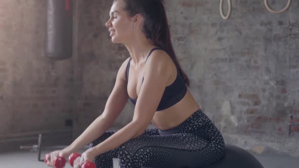 Woman in black sportswear, sitting on fitness ball and doing exercises with dumbell