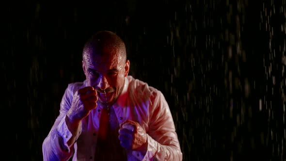 a Man in an Unbuttoned White Shirt Raises Head and Fists and Starts Boxing in Streams of Water on a
