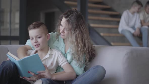 Portrait of Pretty Young Mother Reading Book with Her Son Sitting on the Sofa While Her Two Teen