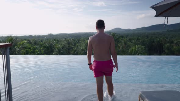 Traveler Man Walks Up Towards Swimming Pool in Swim Shorts Watching Amazing Tropical Forest Panorama