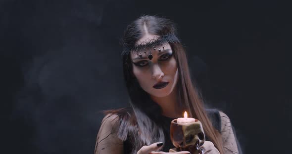 Mystical Female in Black Attire Holds a Skull with a Burning Candle and Looking at the Camera, the