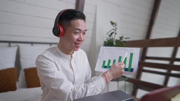 Smiling Chinese Young Man in Headphones Pointing at Graph Talking Using Virtual Conference on Laptop