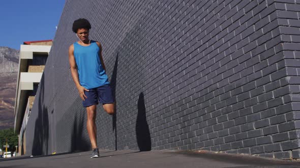 Fit african american man exercising in city, using earphones, stretching legs in street