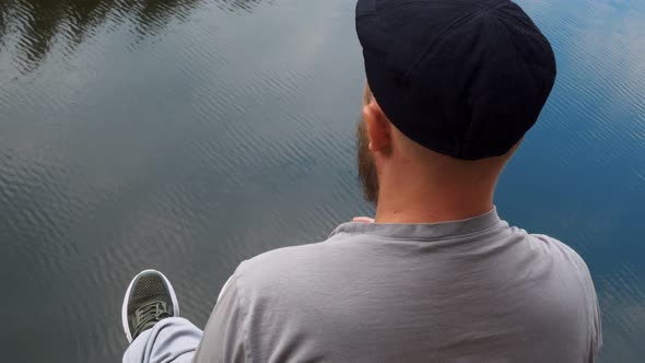 A Man in a Cap with a Beard is Sitting on a Cliff and Throwing Stones Into the Water From a Height