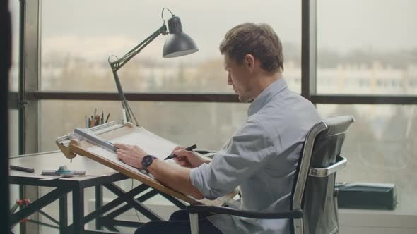 A Male Architect Sitting at a Table Draws a Plan of the Building and is Engaged in Design