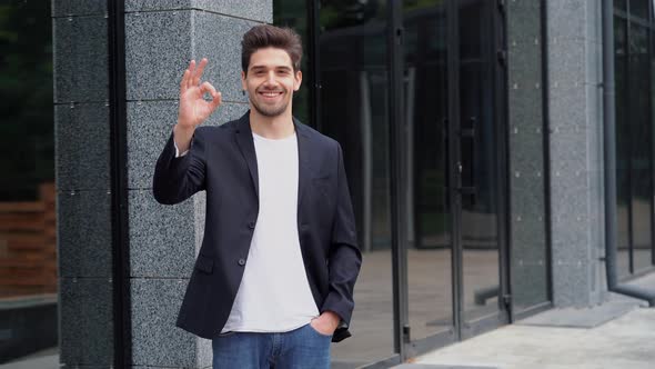 Successful happy man looking to camera and showing OK sign on office building background. 