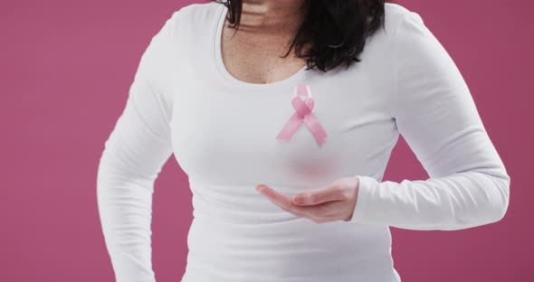 Mid section of woman with pink ribbon on her chest holding invisible object against pink background