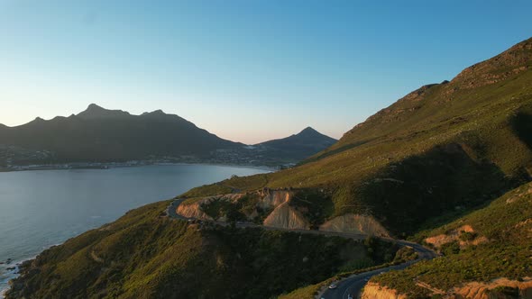 Cars driving on Chapmans Peak Road at sunset with Hout Bay in background, aerial