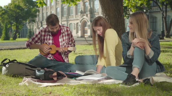 Positive Caucasian Man Playing Ukulele for Beautiful Girls on Sunny Green Meadow. Portrait of Young