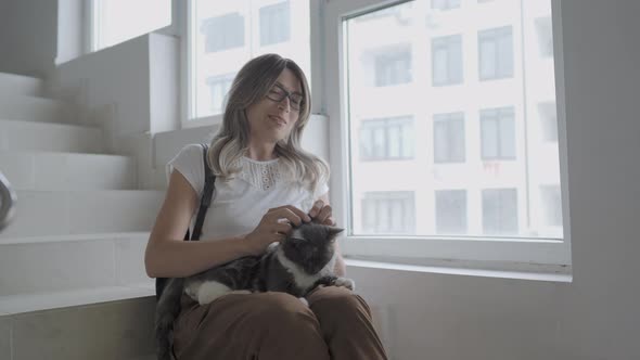 Pretty Woman Strokes Colorful Stray Cat While Sitting on White Steps of Staircase of Apartment