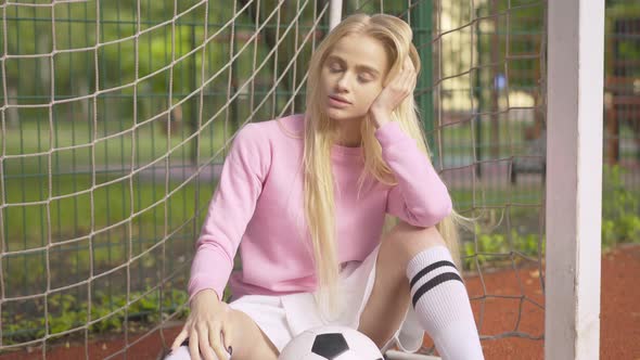 Charming Young Blond Woman Sitting with Football Ball on Outdoor Sports Ground. Camera Approaches To