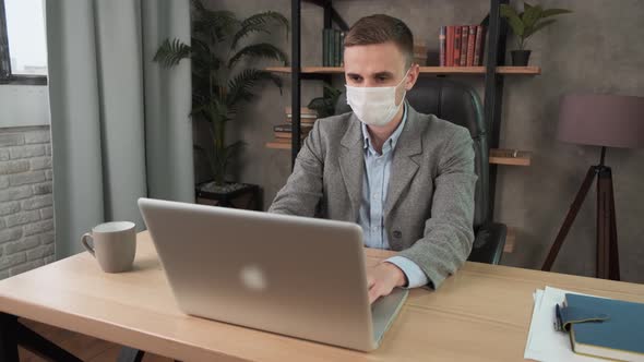 Young Confident Man Employee in a Medical Facial Mask Works in the Office at the Computer. Male