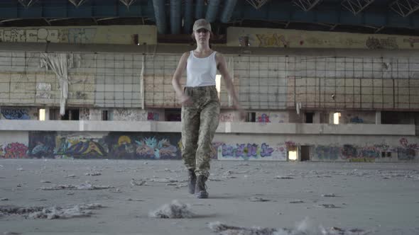Attractive Young Woman in Military Uniform Marching on Concrete Floor in Dusty Dirty Abandoned Place