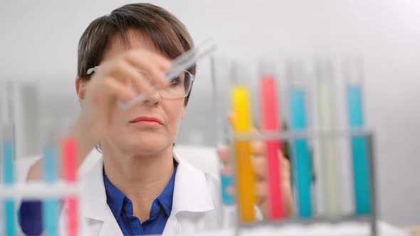 Focused Mature Female Chemist in Safety Glasses Mixing Colorful Reagent Tube Making Researching