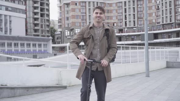 Portrait of Cheerful Adult Caucasian Man Standing on City Street with Scooter and Smiling at Camera