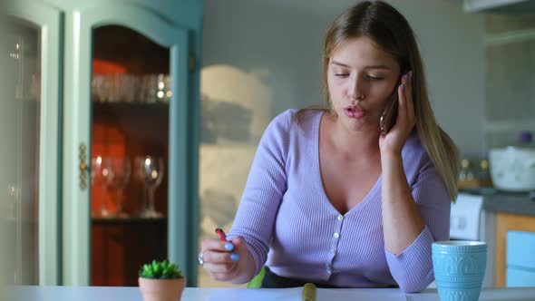 Young European Woman is Talking on Phone and Writing at Table in Apartment Room Rbbro