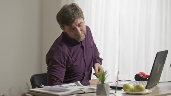 Middle Shot of Sad Tired Man with Backache Warming Up Sitting at Table with Laptop