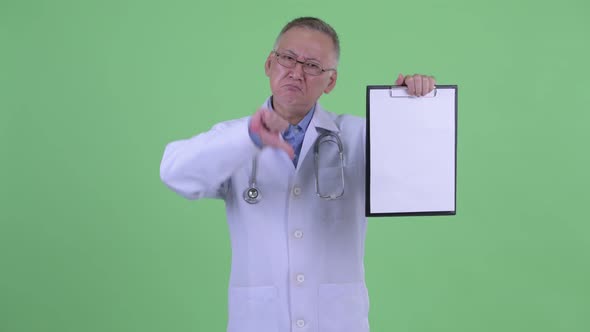 Stressed Mature Japanese Man Doctor Showing Clipboard and Giving Thumbs Down