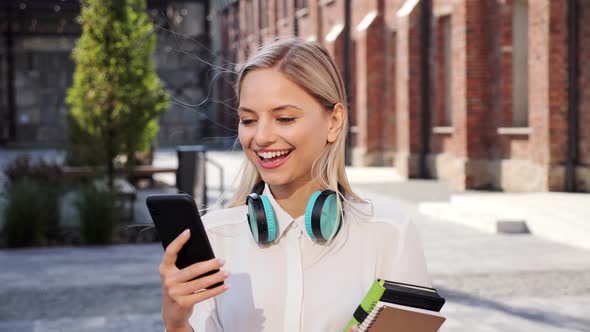 Attractive Young Girl Student with Headphones Holding Books While Use Phone