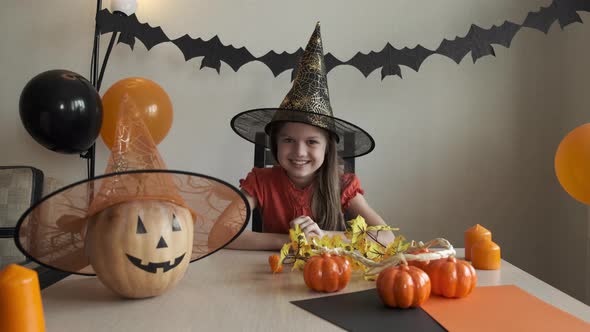 Cute Little Girl Wearing Witch Hat Sitting Behind a Table in Halloween Theme Decorated Room Looking