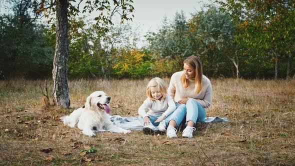 Young Mother is Smiling and Talking to Her Little Daughter While Sitting on Blanket at Autumn Park