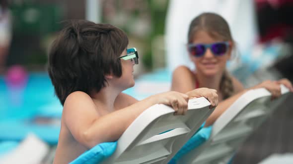 Cute Caucasian Boy in Sunglasses Posing on Sunbed with Blurred Sister Smiling at Background