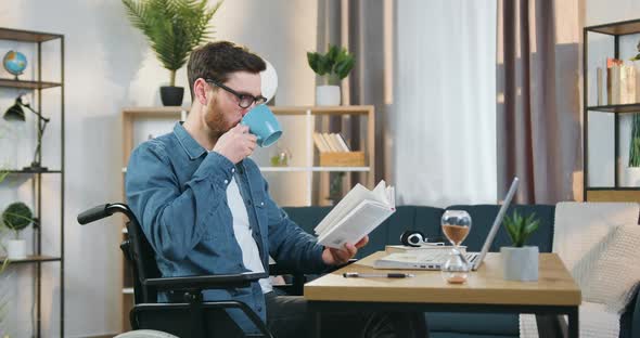 Man in Glasses Sitting in Wheelchair and Reading Book Simultaneously Drinking Tea at Home