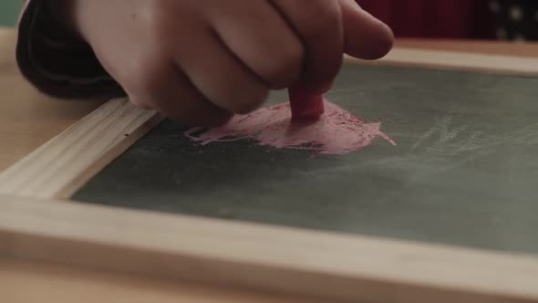 A child draws a pink heart on a slate with chalk as the camera tracks right.