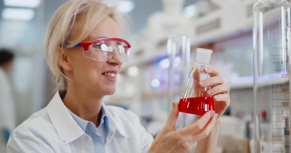 Smiling Female Scientist in Goggles with Chemicals in Flask Making Test or Research in Laboratory
