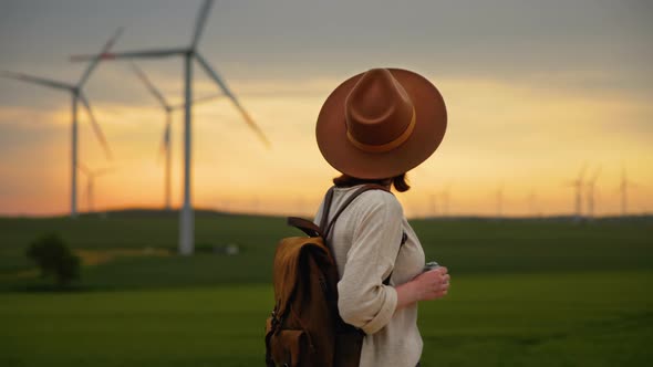 Young photographer in a hat and with a backpack looking at a wind farm in a field