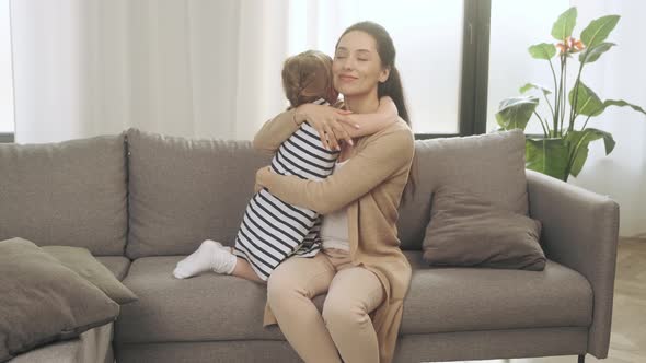A Young Mother Gently Hugs Her Little Daughter Sitting on the Couch at Home