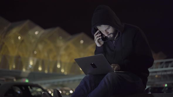 Male Hacker in Hood Talking on Phone and Teleworking on Laptop