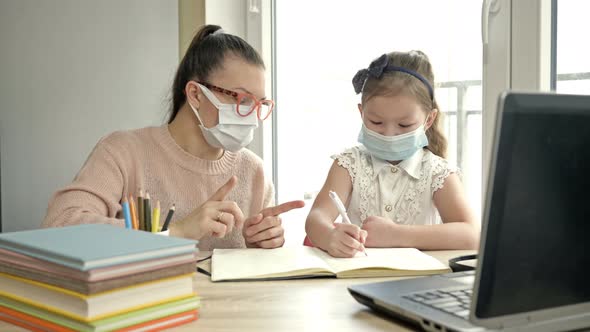 Mom or Teacher or Tutor Helps an Elementary School Student with Her Lessons. Both Have Medical Masks