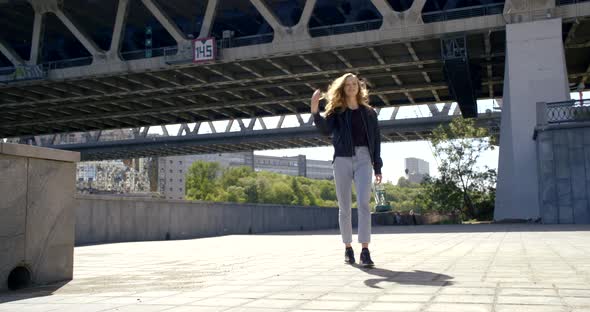 A Beautiful Slender Girl in Jeans and a Leather Jacket Walks Under a High Bridge Against a Stone