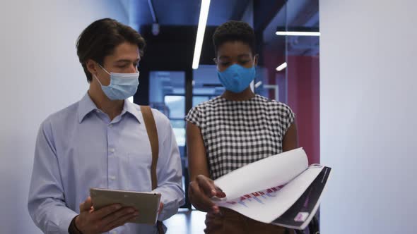 Diverse business colleagues wearing face masks using tablet in discussion walking through office