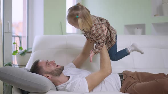 Side View of Cheerful Young Father Having Fun Holding Baby Girl Up Imitating Airplane As Joyful