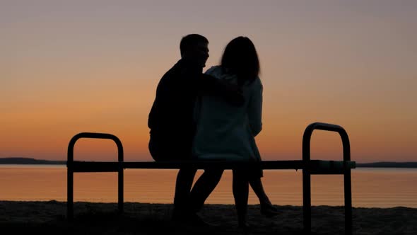 Loving Couple Sitting On A Bench In An Embrace On The Beach And Enjoy The Sunset