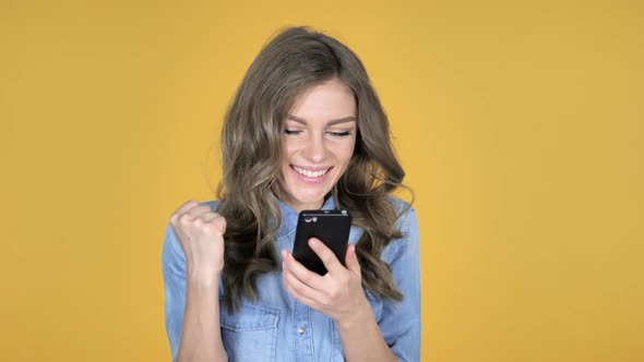 Young Girl Excited for Success While Using Smartphone Isolated on Yellow Background