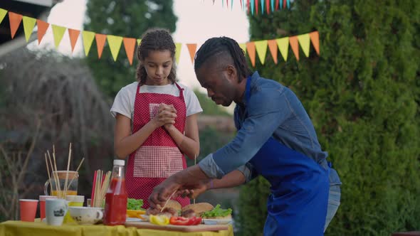 Joyful Teenage Daughter Clapping and Helping Serious Father Preparing Picnic Burgers Outdoors