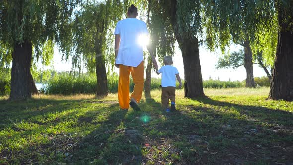 Young Father and Little Son Holding Hands and Walking Through Green Park at Summer Day