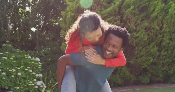 Happy biracial man holding piggyback his fiance with engagement ring on hand in garden in sun