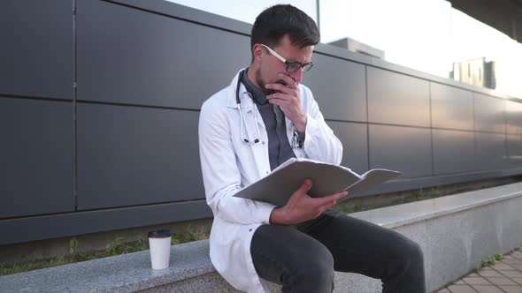 Young Male Doctor Works with Medical Documents During Coffee Break in Backyard Hospital. Tired