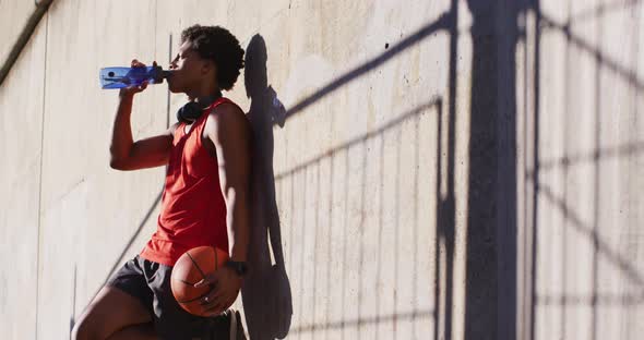 Fit african american man exercising in city taking a break, drinking water, holding basketball
