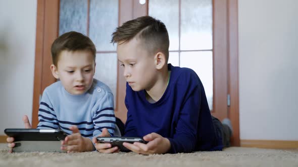 Cute boys lying on the floor in the living-room and playing on modern devices with excited faces.