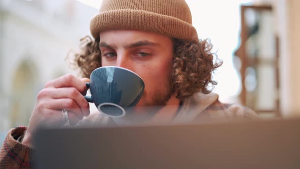 Confident curly-haired man looking at laptop and drinking coffee in cafe