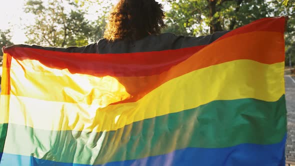 Afroamerican Lady is Holding Multicolored LGBT Pride Flag and Waving It Looking Back and Smiling