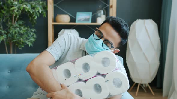 Portrait of Arab Man in Medical Mask Holding Pack of Toilet Paper Lying on Couch at Home