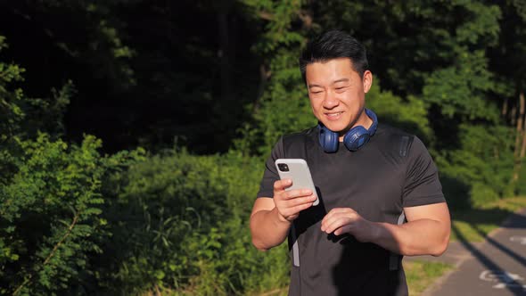 Happy Asian Man Use Smartphone App for Sports Results with Headphone in Park